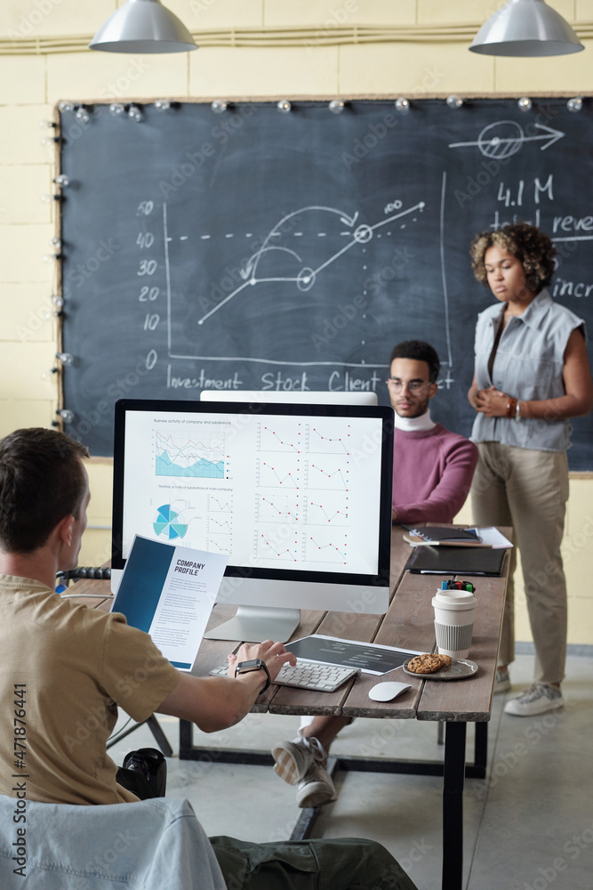 Poster Young economist analyzing graphs on computer screen on background of two biracial colleagues working in front of monitor