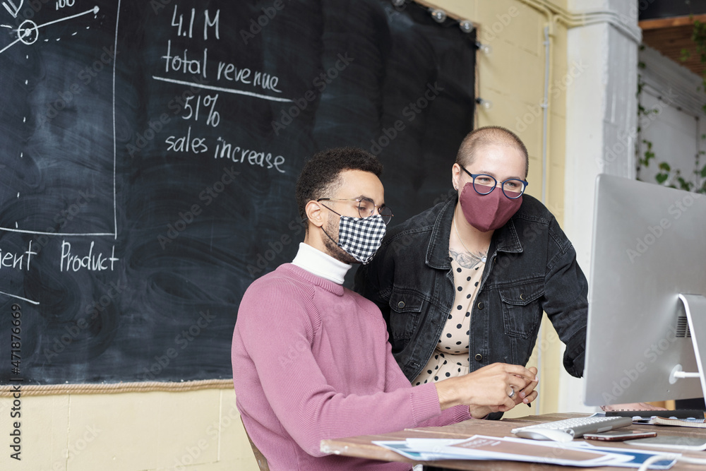 Poster Two young office workers in protective masks looking at computer screen during discussion of financial data at meeting