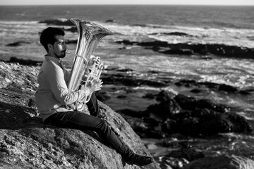 Musician sitting on the rocks with a tuba on the ocean shore. Black and white photo..