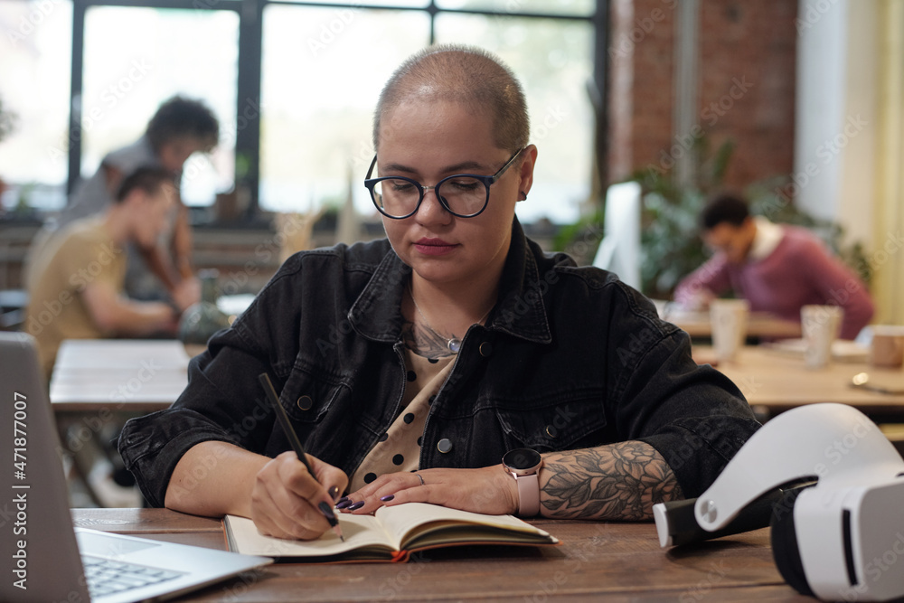 Poster young serious businesswoman making notes in notebook while sitting by workplace in front of laptop w
