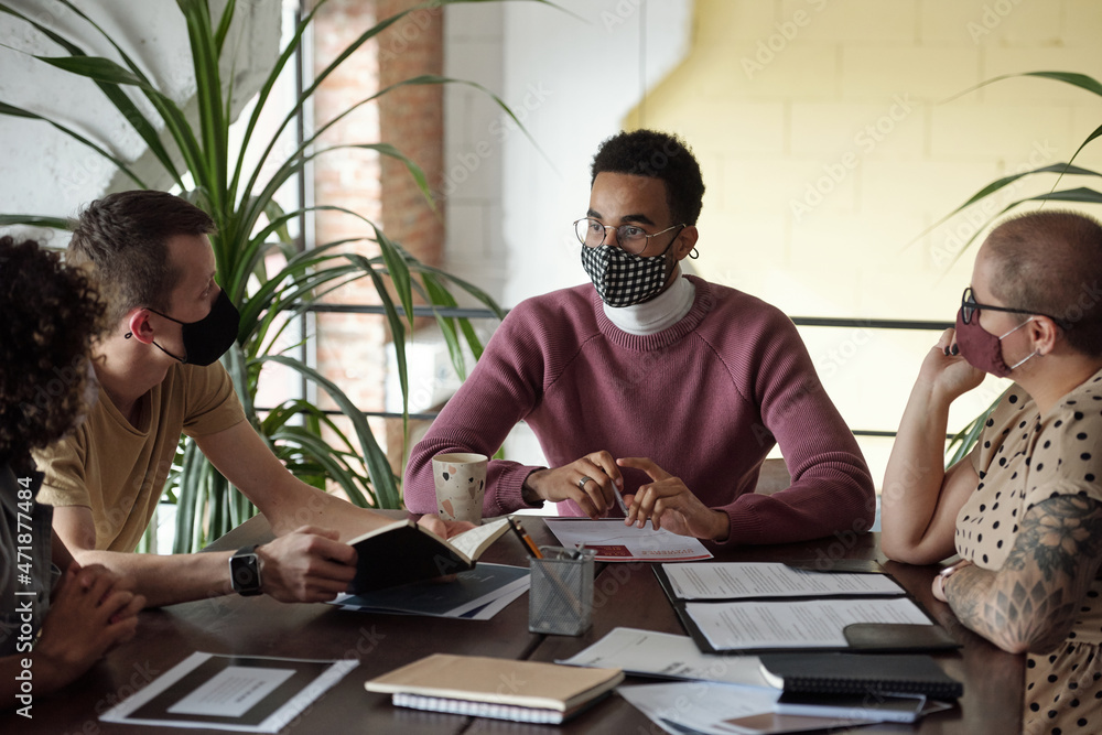 Poster young confident businessman in mask sitting by table in front of co-workers during discussion of wor