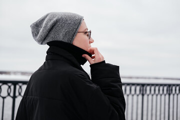 Pensive thoughtful young woman with short hair in hat, eyeglasses, black coat walking on a winter...