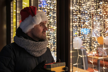 Man in Santas hat looking and dreaming in illuminated shop window. Christmas holidays sales concept