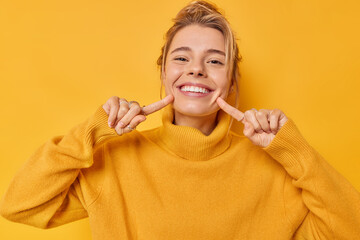 Portrait of cheerful young woman smiles broadly indicates index fingers at face wears casual jumper expresses positive emotions isolated over vivid yellow background. Look at my brilliant smile - 471879400