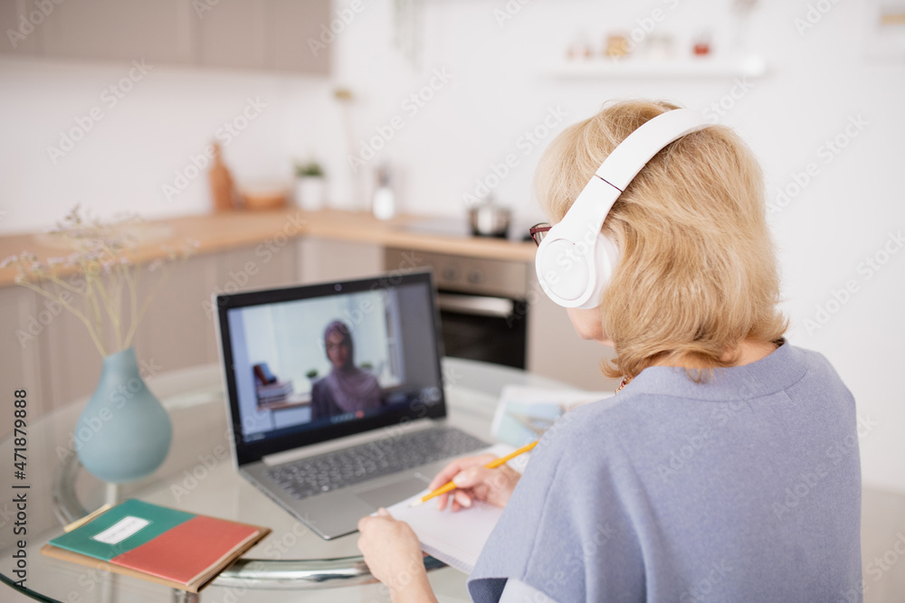 Poster back view of mature female student making notes in front of laptop while teacher on screen explainin