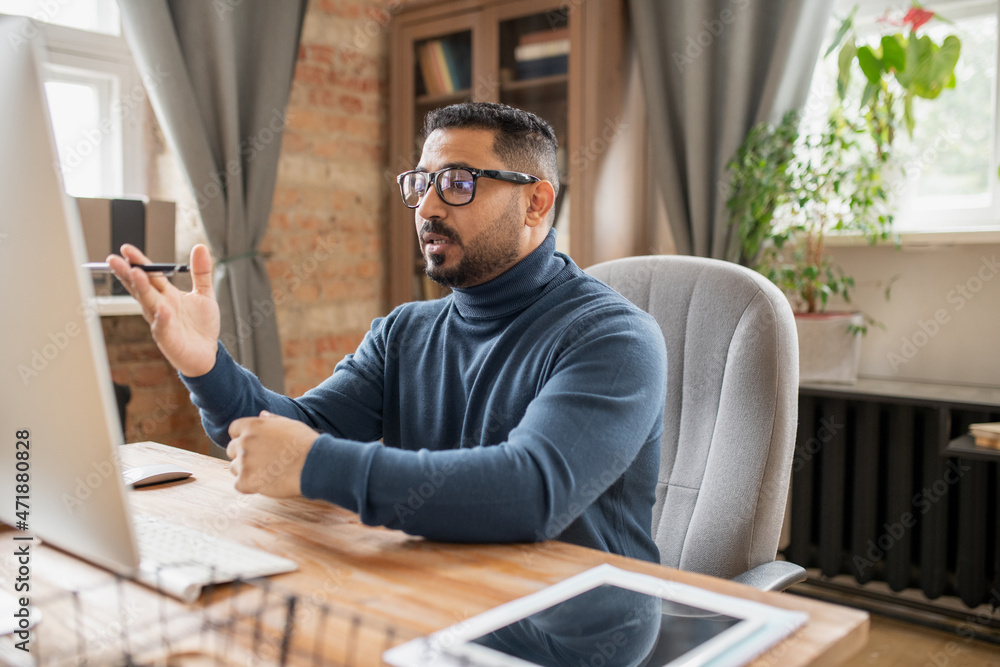 Poster confident middle aged teacher explaining something to online audience while sitting in front of comp