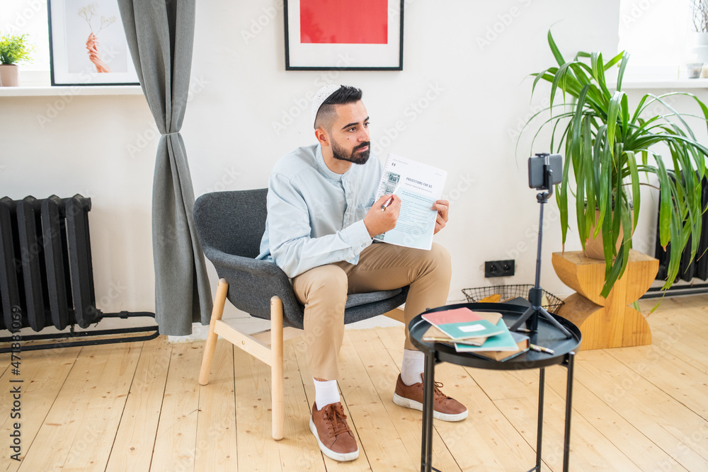 Aufkleber young arabic man pointing at paper while sitting in armchair in front of smartphone camera during on