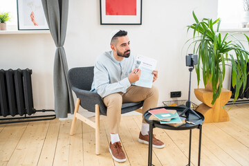 Young Arabic man pointing at paper while sitting in armchair in front of smartphone camera during online lesson or training