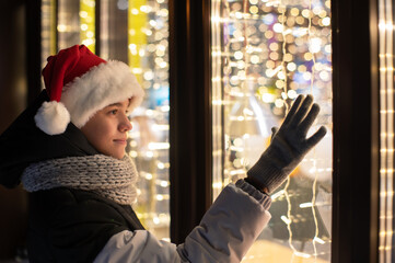 Boy in Santas hat looking and dreaming in illuminated shop window. Xmas presents holidays, or shopping on New Year or Christmas concept