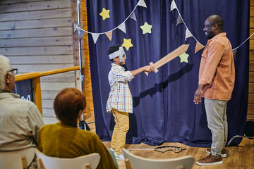 Happy boy in costume of knight holding sword against his father during performance in front of...