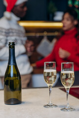 Wineglasses with champagne and bottle on table against blurry happy couple in festive knitted sweaters communicating in Christmas atmosphere