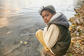 Cute youthful boy in warm casualwear sitting on squats by waterside during chill on autumn day and...