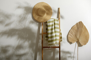 Organic linen beach towel on wood ladder, straw hat and dried palm leaf against white wall with...