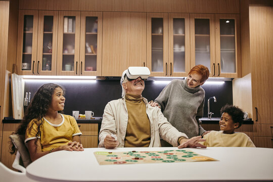 Two cute interracial siblings and their grandmother looking at mature man in vr headset while playing board game together