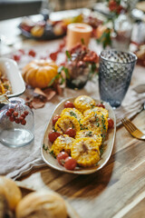 Plate with baked chopped corn and fresh cherry tomatoes standing among kitchenware and decorations on wooden dinner table