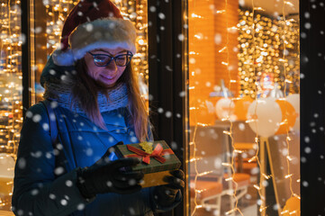 Pretty Woman in Santas hat with gifts box near illuminated cafe window. She waves her hand and waits for her beloved partner. Xmas presents holidays, or shopping on New Year or Christmas concept