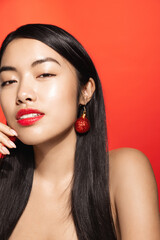Fototapeta premium Vertical portrait of sexy, sensual asian woman wears christmas earrings, xmas tree decor in ear, gazing sensual at camera, red background
