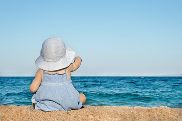 Fototapeta na wymiar Baby girl wearing sunhat,sitting on sandy beach,look at sea,shot from back.Copy space