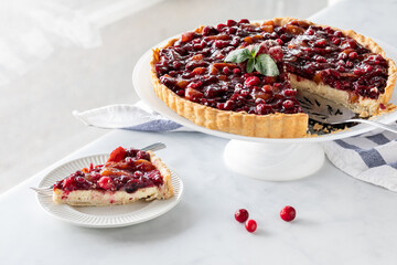 A cranberry tart with one slice on a plate, against a bright window.