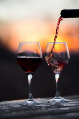 Two wine glasses, one of the glasses is full of red wine and another one is being poured, with a sunset background