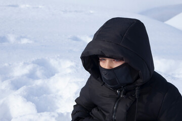 A young boy in warm black clothes with tears in his eyes from the cold against the background of white snow.