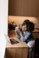  Young african lady is relaxing at home with cup of tea and reading e-book on laptop while enjoying calm indoor lifestyle. Girl with short dark curly hair happy. Freelancer, networking, site concept.