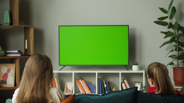 Women watching television with chroma green screen. Girlfriends switching channels on tv with remote controller, sitting on sofa together in living room. Spare time at home, relaxing.