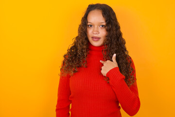 Teenager girl wearing red sweater over yellow background being in stupor shocked, has astonished expression pointing at oneself with finger saying: Who me?