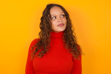 Teenager girl with curly hair wearing red sweater over yellow background, looks pensively aside, plans actions after university, imagines what to do Thinks over about new project.