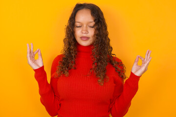 Teenager girl with curly hair wearing red sweater over yellow background doing yoga, keeping eyes...