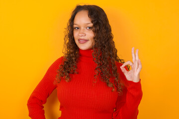 Glad attractive Teenager girl with curly hair wearing red sweater over yellow background shows ok sign with hand as expresses approval, has cheerful expression, being optimistic.