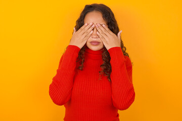 Teenager girl with curly hair wearing red sweater over yellow background covering eyes with both hands, doesn't want to see anything or feeling ashamed. Human feelings reactions.