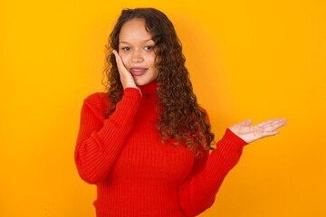 Positive glad teenager girl wearing red knitted sweater against yellow background says: wow how exciting it is, indicates something.  One hand on his head and pointing with other hand.