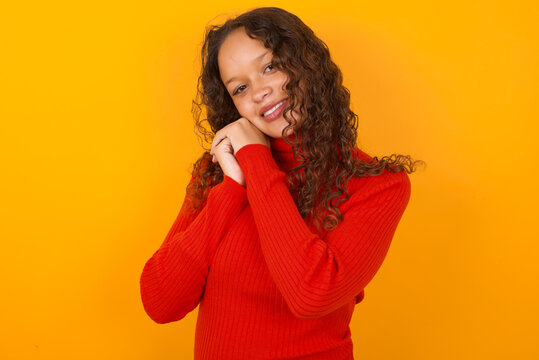 Dreamy Teenager girl with curly hair wearing red sweater over yellow background with pleasant expression, closes eyes, keeps hands crossed near face, thinks about something pleasant