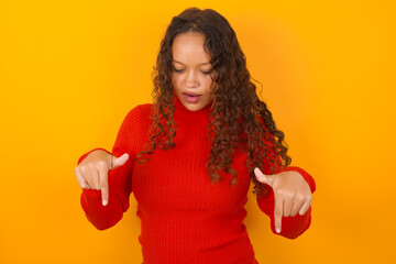Amazed teenager girl wearing red knitted sweater against yellow background points down with fore...