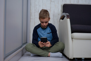 młody chłopak siedzi na podłodze między fotelem a szafą używa telefonu  - obrazy, fototapety, plakaty