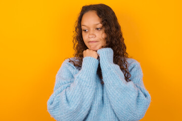 Curious Teenager girl wearing blue sweater over yellow background keeps hands under chin bites lips and looks with interest aside.