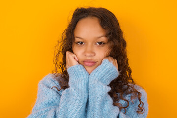 Portrait of sad Teenager girl wearing blue sweater over yellow background hands face