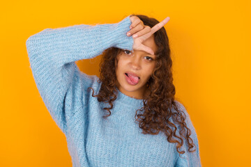 Funny Teenager girl wearing blue sweater over yellow background makes loser gesture mocking at...