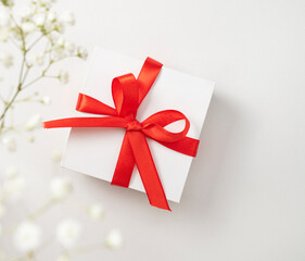 a white gift box with a red ribbon on a white  background with a bouquet of  gypsophila. The concept of a wedding or gift card. Top  view and copy space