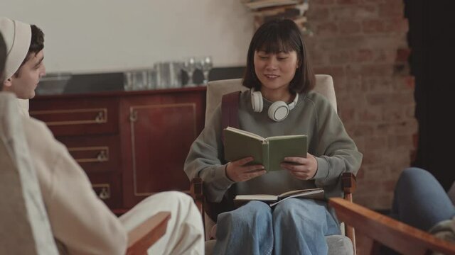 Handheld medium slowmo shot of female Asian student reading book aloud sitting in circle together with other students during Literary club meeting at campus
