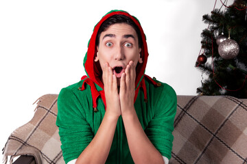 A handsome man dressed as Santa's helper sits on the couch with a surprised face. White background...