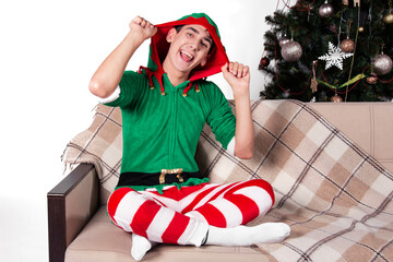A handsome man dressed as Santa's helper sits on the couch with a surprised face. White background...