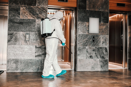 Air cleaning and disinfection of the space in the hotel lobby. A professional of the unknown sex gender refreshes the passage to the elevator while carrying a fluorescent yellow tank on his back
