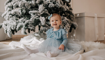 a baby girl in a blue festive dress sits on a white plaid near the Christmas tree and looks into the camera