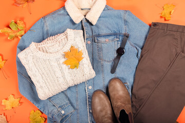 Denim jacket, sweater, smartwatch, pants, shoes and autumn leaves on color background, closeup
