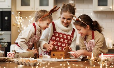 Happy family mother and two kids in xmas aprons decorating Christmas honey gingerbread house - obrazy, fototapety, plakaty