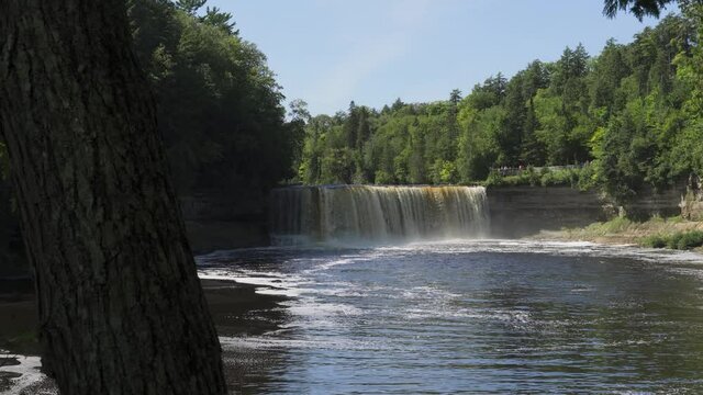 Long View of Higher Falls of Tahquamenon Falls; rushing water drops into pool below, trees frame the banks; low angle.