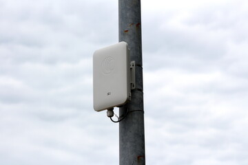Modern new wireless communication device in white plastic case tied with plastic ties to old metal utility pole with rusted patches on dark gloomy cloudy sky background