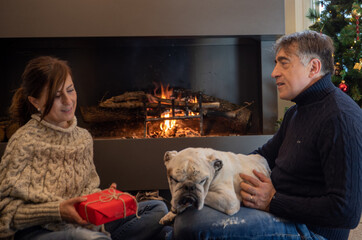 Middle-aged couple exchanges Christmas presents in front of the fireplace with their dog in the living room of the house.